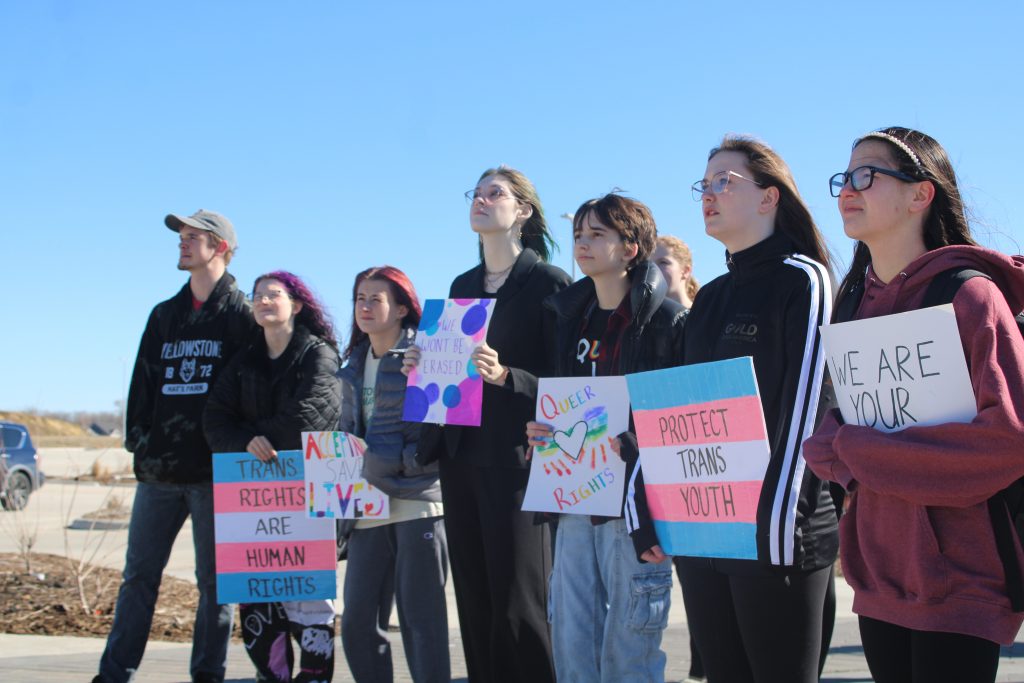 students stand with signs that read "trans rights are human rights", "queer rights", and "protect trans youth"